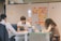 Three individuals are focused on their laptops at a table in an office setting, with a whiteboard covered in colorful sticky notes in the background. The room has a collaborative and concentrated atmosphere.