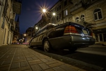 A nighttime street scene with a black luxury sedan parked on the side of a dimly-lit city road. Surrounding buildings are older with classic architecture, and streetlights cast a warm glow, creating reflections on the car's surface. The sky is dark, emphasizing the lights and creating an urban atmosphere.