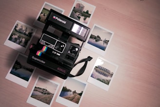 A vintage Polaroid camera is surrounded by several instant photographs depicting various outdoor scenes such as buildings, a bridge, and a landscape, all placed on a light wooden surface. The camera is the focal point, with a colorful stripe decal, and the photos appear slightly scattered.