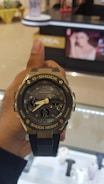 A hand holds a G-Shock wristwatch with a metallic and black design, featuring analog hands and a digital display showing the time and day. In the background, there is a blurred image of a store with a poster displaying a woman's face.