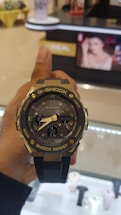 A hand holds a G-Shock wristwatch with a metallic and black design, featuring analog hands and a digital display showing the time and day. In the background, there is a blurred image of a store with a poster displaying a woman's face.