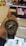 A hand holds a G-Shock wristwatch with a metallic and black design, featuring analog hands and a digital display showing the time and day. In the background, there is a blurred image of a store with a poster displaying a woman's face.