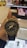 A hand holds a G-Shock wristwatch with a metallic and black design, featuring analog hands and a digital display showing the time and day. In the background, there is a blurred image of a store with a poster displaying a woman's face.