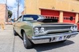 A classic car is parked in front of a building with red garage doors. The vehicle is an older model, possibly a muscle car, featuring a two-tone paint job with a black hood and a light green body. Its chrome fixtures and vintage styling emphasize the era of its design. The surrounding area is an urban setting with brick buildings and utility poles visible in the background.