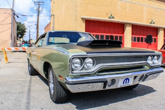 A classic car is parked in front of a building with red garage doors. The vehicle is an older model, possibly a muscle car, featuring a two-tone paint job with a black hood and a light green body. Its chrome fixtures and vintage styling emphasize the era of its design. The surrounding area is an urban setting with brick buildings and utility poles visible in the background.