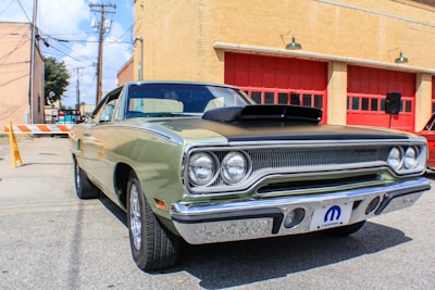 A classic car is parked in front of a building with red garage doors. The vehicle is an older model, possibly a muscle car, featuring a two-tone paint job with a black hood and a light green body. Its chrome fixtures and vintage styling emphasize the era of its design. The surrounding area is an urban setting with brick buildings and utility poles visible in the background.