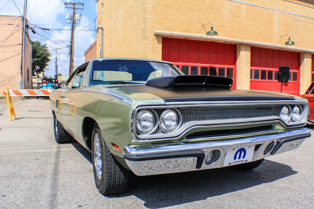 A classic car is parked in front of a building with red garage doors. The vehicle is an older model, possibly a muscle car, featuring a two-tone paint job with a black hood and a light green body. Its chrome fixtures and vintage styling emphasize the era of its design. The surrounding area is an urban setting with brick buildings and utility poles visible in the background.
