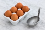 A carton of six brown eggs is placed on a white marble surface. Next to the carton, there is a metal egg poacher with holes at the bottom and a handle.