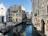 A picturesque canal scene with gondolas floating gently through the water, bordered by historic buildings with ornate facades and vibrant colors. People are walking along the waterfront, enhancing the lively atmosphere. A small arched bridge in the background adds charm to the setting.