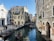 A picturesque canal scene with gondolas floating gently through the water, bordered by historic buildings with ornate facades and vibrant colors. People are walking along the waterfront, enhancing the lively atmosphere. A small arched bridge in the background adds charm to the setting.