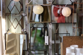 Several rectangular and diamond-shaped mirrors are displayed, hanging on a rustic wooden backdrop. Reflections include string lights and a person in a red coat. Some price tags are visible, and a few white cylindrical objects are in the foreground.