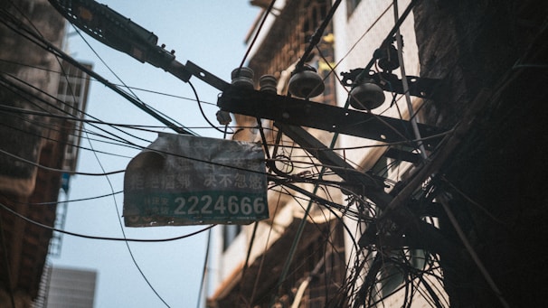 An entanglement of electric wires is attached to a pole mounted with several ceramic insulators, creating a complex web of cables. A weathered street sign with the number 2224666 is visible, and the scene is set against the backdrop of aged urban buildings.