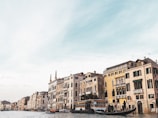 A scenic canal side with historical buildings displaying classical architectural styles. Gondolas and a boat with people are on the water, indicating a lively and traditional transport scene unique to this region.