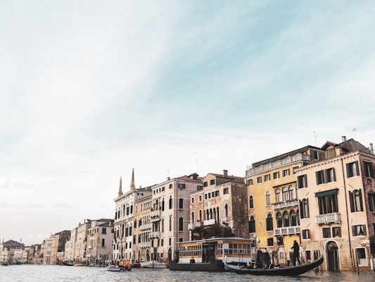 A scenic canal side with historical buildings displaying classical architectural styles. Gondolas and a boat with people are on the water, indicating a lively and traditional transport scene unique to this region.