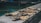 Several dried fish are laid out in a row on a concrete surface near what appears to be a body of water in a harbor or dock area. The fish are secured with strings. The background shows blurred boats and the water reflects a greenish hue.