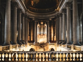 The grand interior of a classical architecture building features tall, ornate columns lining the sides, with a high arched ceiling decorated with intricate paintings. A lavish organ is positioned at the far end, surrounded by elaborate carvings and gold accents. Warm lighting highlights the details of the decoration.