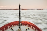 The image features the front of a ship navigating through an icy body of water. The water appears partially frozen, creating a textured pattern on its surface. The ship's bow is painted bright red, providing a striking contrast against the pale ice. In the background, there are small landmasses with sparse vegetation under a cloudy sky.