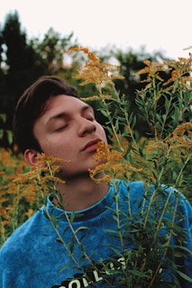A young person stands amidst tall green plants with yellow flowers. Their eyes are gently closed, and they appear to be at peace, enjoying the natural surroundings. The sky is overcast, adding a soft light to the scene. The person is wearing a blue shirt.