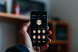 A hand holds a smartphone displaying a screen where various cartoon emoji faces are featured. The background is softly blurred, indicating an indoor setting with possibly a shelf or furniture.