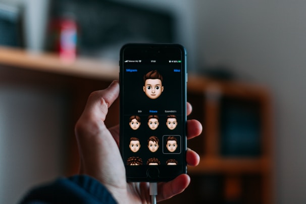 A hand holds a smartphone displaying a screen where various cartoon emoji faces are featured. The background is softly blurred, indicating an indoor setting with possibly a shelf or furniture.