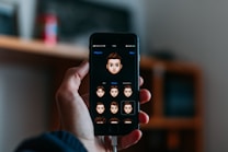 A hand holds a smartphone displaying a screen where various cartoon emoji faces are featured. The background is softly blurred, indicating an indoor setting with possibly a shelf or furniture.