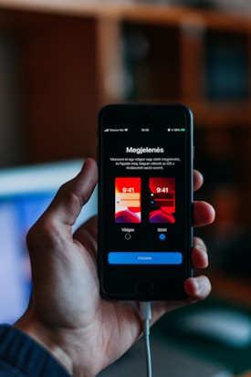 A hand holding a smartphone that displays a screen with options for selecting a light or dark mode in the Hungarian language. The phone shows two images of user interface themes, with options to choose between them. The background is softly blurred, suggesting a home or office environment.