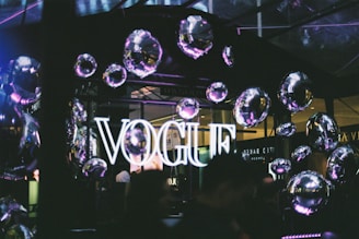 A neon sign displaying the word 'VOGUE' is illuminated against a backdrop of reflective, metallic balloons that create a festive atmosphere. The setting appears to be indoors with a dark, ambient lighting, enhancing the glow of the sign and the shiny surfaces of the balloons.