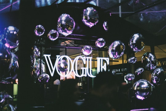 A neon sign displaying the word 'VOGUE' is illuminated against a backdrop of reflective, metallic balloons that create a festive atmosphere. The setting appears to be indoors with a dark, ambient lighting, enhancing the glow of the sign and the shiny surfaces of the balloons.