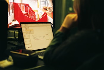 A person is sitting at a desk and looking at a computer screen displaying a red architectural design. Next to it, a laptop shows a mind map. The setting appears to be an office or workspace with a focus on creative tasks.