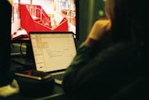 A person is sitting at a desk and looking at a computer screen displaying a red architectural design. Next to it, a laptop shows a mind map. The setting appears to be an office or workspace with a focus on creative tasks.