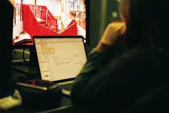 A person is sitting at a desk and looking at a computer screen displaying a red architectural design. Next to it, a laptop shows a mind map. The setting appears to be an office or workspace with a focus on creative tasks.