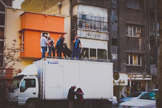 A group of people stands on top of a white truck parked on a street, surrounded by urban buildings. Some individuals appear to be setting up filming equipment. The scene takes place in a busy city area with a mixture of old and new architecture.