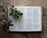 An open book displaying a passage from the Bible with a branch of green leaves and acorns resting on it. The book is placed on a rustic wooden surface, and the pages are gently curving under the branch's weight.