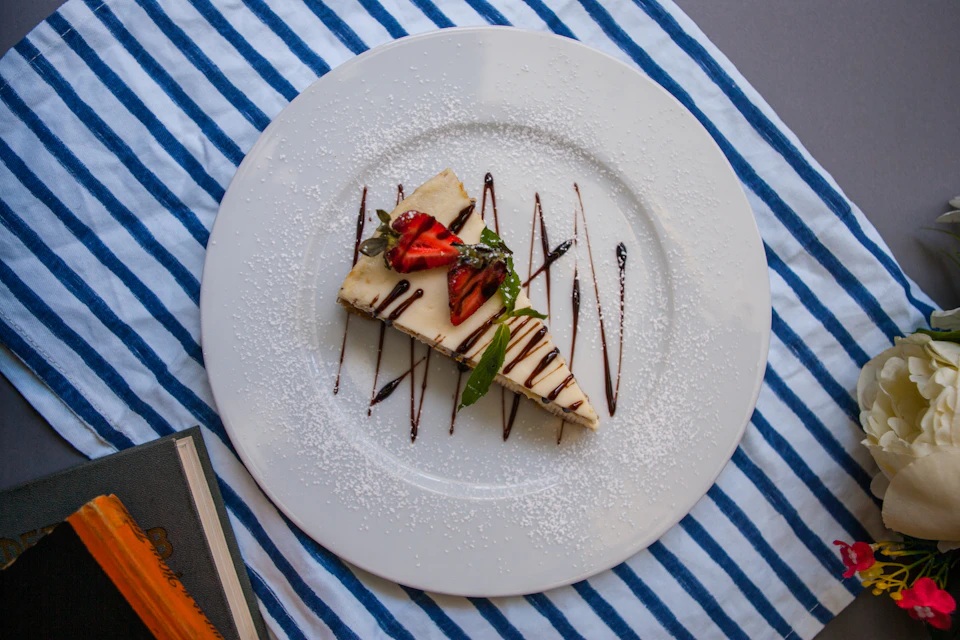 The image shows a slice of cake on a white plate with a blue rim, placed on a table covered with a striped tablecloth. The cake is adorned with chocolate sauce and fresh strawberries, adding to its visual appeal. A book can be seen in the background, suggesting that this might be a cozy reading or dining experience.