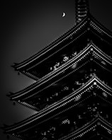 A dark, moody black and white photograph captures the silhouette of a traditional multi-tiered pagoda with intricate architectural details. A crescent moon appears in the sky above, adding an ethereal quality to the scene.