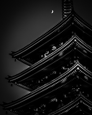 A dark, moody black and white photograph captures the silhouette of a traditional multi-tiered pagoda with intricate architectural details. A crescent moon appears in the sky above, adding an ethereal quality to the scene.