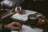 Close-up of hands sketching travel concepts on a notebook beside a steaming cup of coffee.