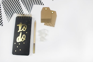 A flat lay photograph of a black spiral notebook with 'to do' in gold lettering on the front cover. Next to it lies a pencil with gold accents, some crystal stones, and several kraft paper gift tags with eyelets. In the background, there are black and white striped triangular party flags.