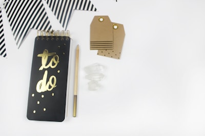 A flat lay photograph of a black spiral notebook with 'to do' in gold lettering on the front cover. Next to it lies a pencil with gold accents, some crystal stones, and several kraft paper gift tags with eyelets. In the background, there are black and white striped triangular party flags.