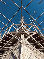 Intricate metal scaffolding and framework extend upwards against a backdrop of clear, blue sky. The structure is composed of intersecting beams and support rods, likely part of a tower or ship’s mast.