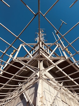 Intricate metal scaffolding and framework extend upwards against a backdrop of clear, blue sky. The structure is composed of intersecting beams and support rods, likely part of a tower or ship’s mast.