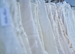 An assortment of wedding dresses on hangers, showcasing various styles and intricate lace details. The array includes multiple layers, elegant embroidery, and differing shades of white and ivory, arranged in a row against a softly lit backdrop.
