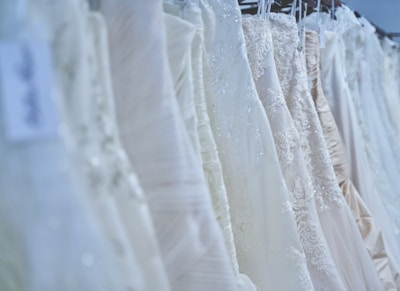 An assortment of wedding dresses on hangers, showcasing various styles and intricate lace details. The array includes multiple layers, elegant embroidery, and differing shades of white and ivory, arranged in a row against a softly lit backdrop.