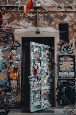 A weathered, urban doorway covered in a variety of colorful stickers and graffiti is set into a stone wall. Above the door, a single black lamp hangs, with patches of exposed brick and various posters visible on the surrounding wall. On the left, a stylized portrait of a person is part of the street art, while a large event poster is pinned to the right side.