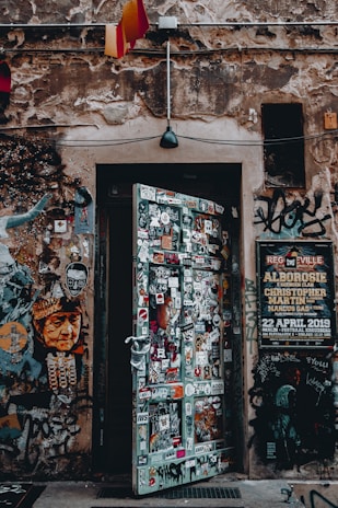A weathered, urban doorway covered in a variety of colorful stickers and graffiti is set into a stone wall. Above the door, a single black lamp hangs, with patches of exposed brick and various posters visible on the surrounding wall. On the left, a stylized portrait of a person is part of the street art, while a large event poster is pinned to the right side.