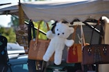 A fluffy white teddy bear is hanging among a collection of leather handbags under a tarp, with a background of trees and a vehicle. The bags vary in color, including brown, tan, and red, and have different styles with embellishments like stitching and buckles.