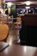 An indoor restaurant setting features wooden chairs and metal bar stools with red seats. The floor is wooden, and in the background there are decorative elements such as a carousel horse and neon lights. The lighting is warm and there are glass partitions with small signs attached to them.
