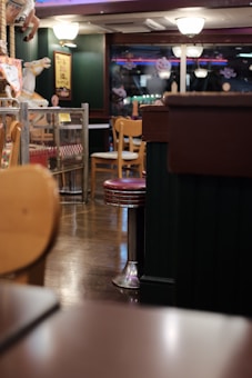 An indoor restaurant setting features wooden chairs and metal bar stools with red seats. The floor is wooden, and in the background there are decorative elements such as a carousel horse and neon lights. The lighting is warm and there are glass partitions with small signs attached to them.