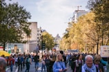 A large group of people are walking down a tree-lined city street, participating in a protest or demonstration. Some hold banners and signs, and there is a sense of movement and engagement among the diverse crowd. City buildings and construction cranes are visible in the background, suggesting an urban setting.