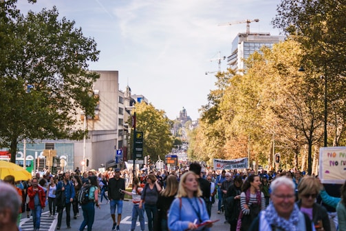 A large group of people are walking down a tree-lined city street, participating in a protest or demonstration. Some hold banners and signs, and there is a sense of movement and engagement among the diverse crowd. City buildings and construction cranes are visible in the background, suggesting an urban setting.